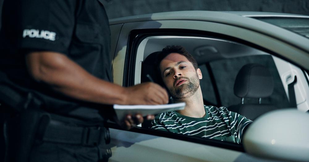Police officer with Police on their sleeve writing on a notepad while speaking to a man seated in a car
