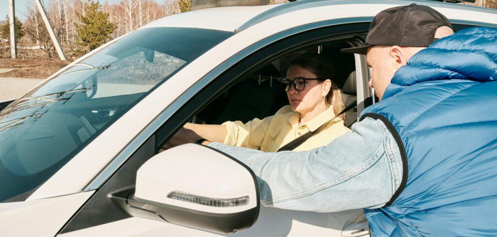 Woman sitting in a car and talking to a man leaning through the window on the driver’s side, suggesting a roadside discussion or possible traffic stop in daylight.