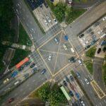An aerial view of a large, busy intersection with multiple lanes of traffic, marked yellow grid boxes, and cars moving and stopping in different directions, surrounded by greenery and adjacent roads.