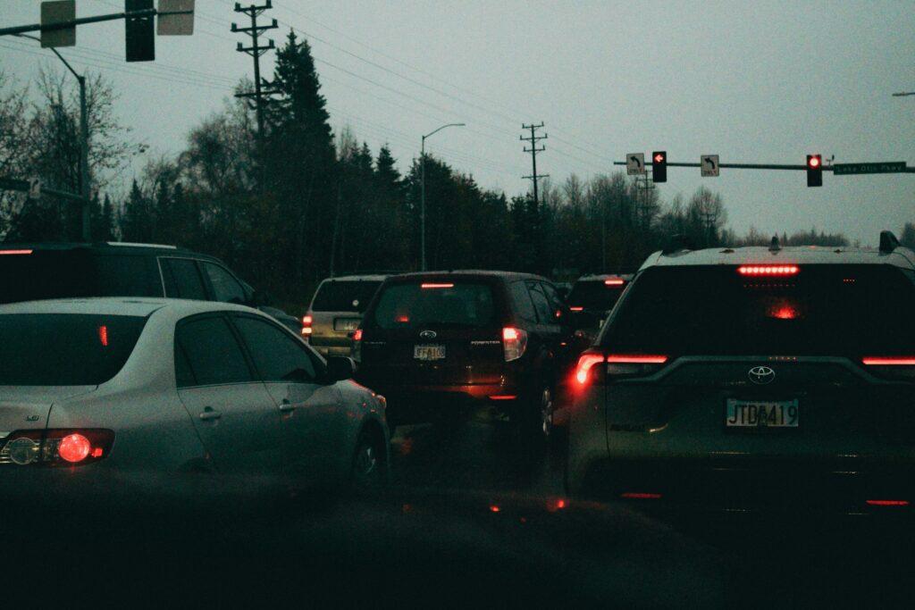 Cars sit in heavy traffic at dusk under a red traffic light, with brake lights glowing and a backdrop of trees, power lines, and an overcast sky creating a dim, crowded roadway scene.