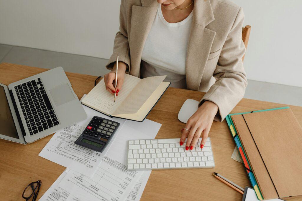 Woman working at a desk with a laptop, calculator, documents, and an open notebook, typing on a keyboard while writing notes.