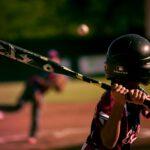 A young baseball player wearing a helmet and gripping a bat prepares to swing as a pitch approaches, with the pitcher blurred in the background on a sunlit field.