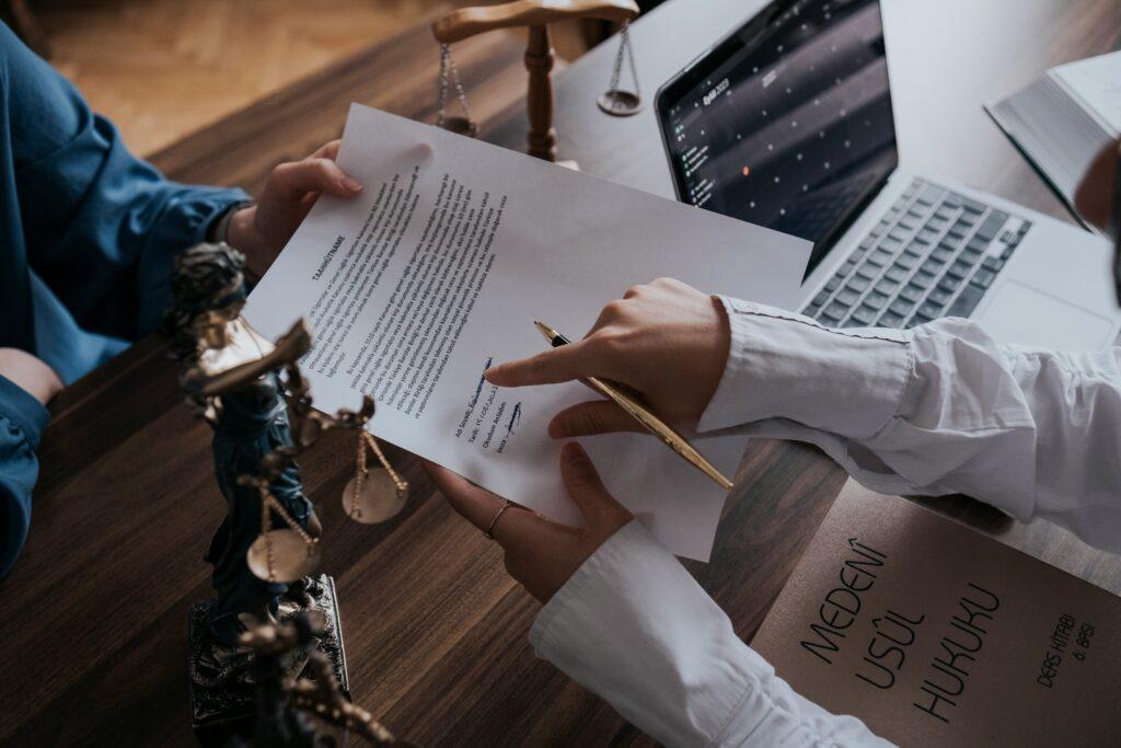 Two people sit at a wooden desk in a law office, reviewing and signing a legal document