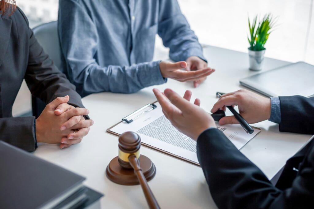 Three people in a professional meeting at a desk with legal documents and a judge’s gavel, discussing a case or agreement in an office setting.