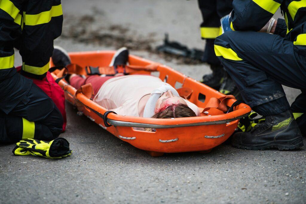 A person with visible head injuries lying on an orange rescue stretcher, secured with straps, while emergency responders in protective gear kneel beside them on the road during a medical rescue situation.