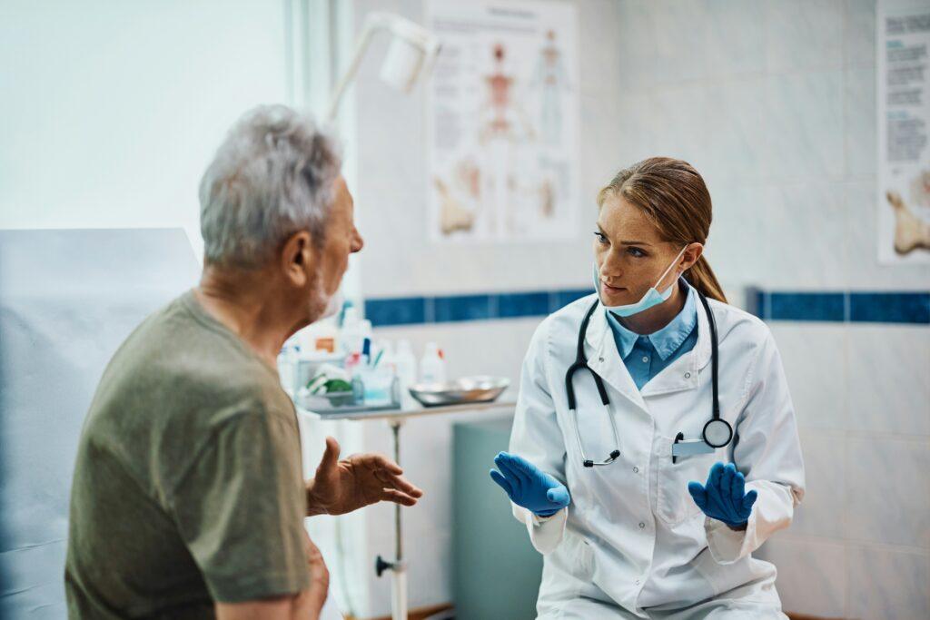 Mature male patient speaking with a female doctor wearing a white coat and stethoscope in a medical examination room, both appearing serious during a discussion about diagnosis or treatment.