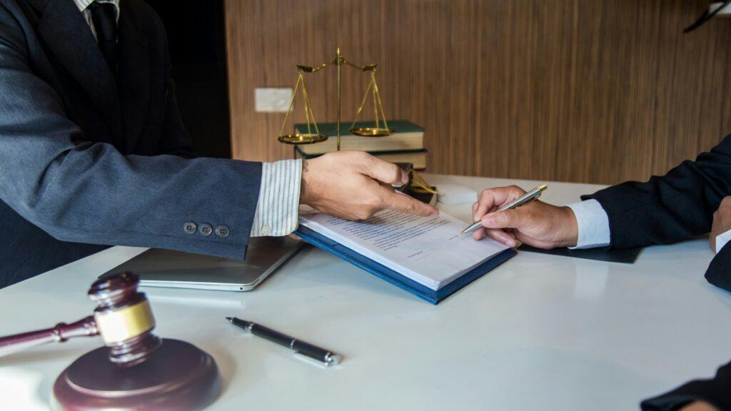 Two people in formal attire sitting at a desk discussing and signing legal documents, with a gavel, scales of justice, and law books visible in the background, symbolizing legal consultation.