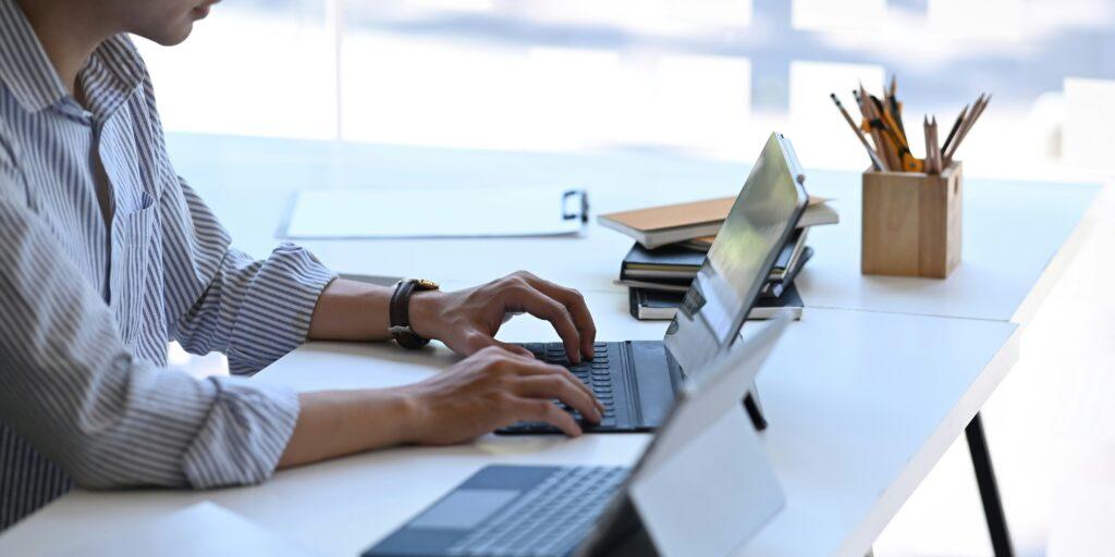 Person working at a desk using a laptop and tablet, surrounded by notebooks and stationery in a bright, modern office setting.