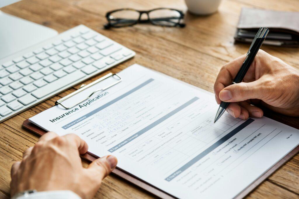 Person filling out an insurance application form on a clipboard with a pen, next to a keyboard, glasses, and a coffee cup on a wooden desk.