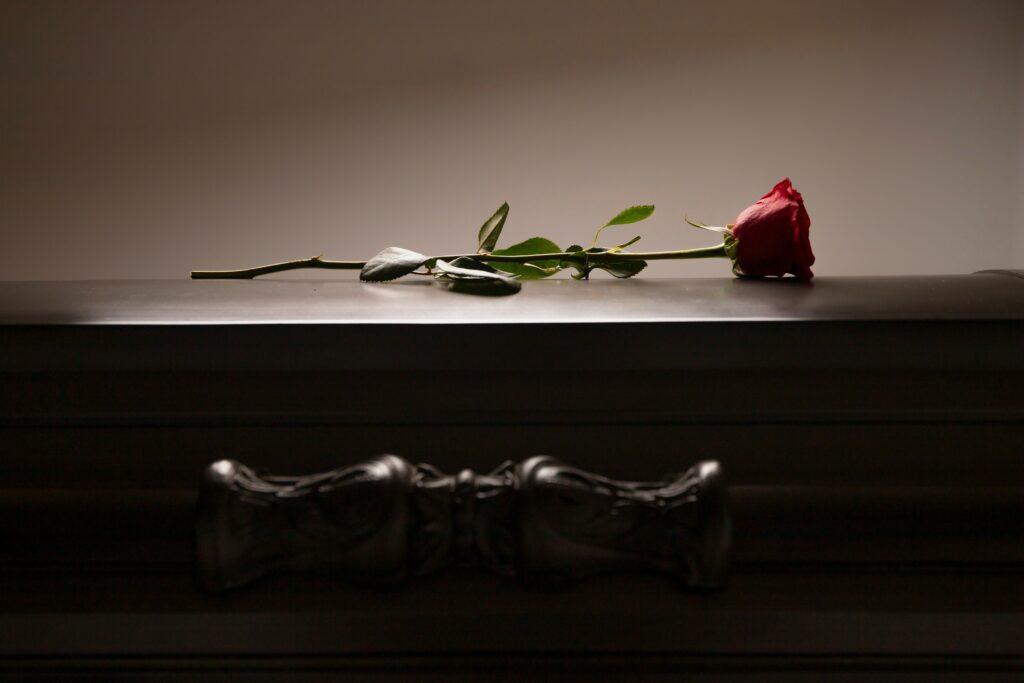 Single red rose resting on top of a closed dark wooden casket in soft, dim lighting.