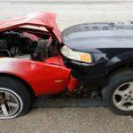 A red car with its hood crushed and engine exposed after a severe front-end collision with a black vehicle, both cars pressed tightly together on the street with visible body and bumper damage.