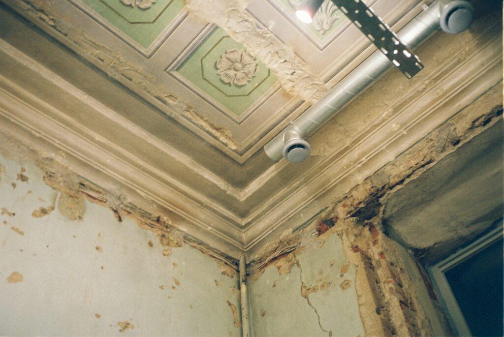 Close-up of a deteriorating apartment ceiling and wall corner with peeling plaster, exposed bricks, and visible water damage, showing signs of long-term neglect and poor maintenance.