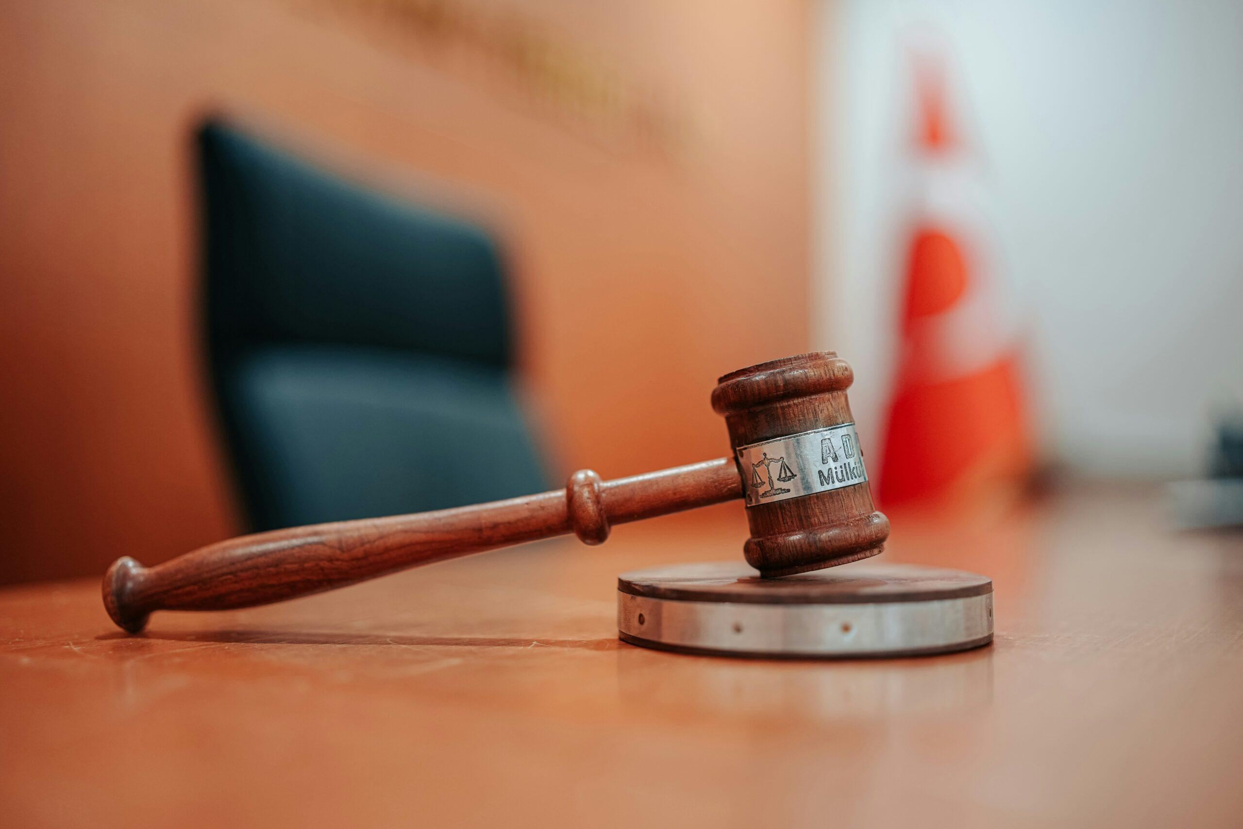 A wooden judge’s gavel rests on a sound block in a courtroom, with a blurred chair and a red-and-white flag visible in the background, symbolizing justice and legal authority.