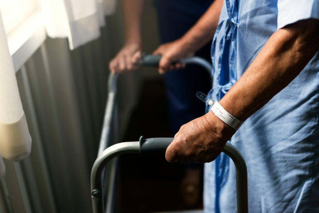 Close-up of a hospital patient wearing a medical gown and wristband using a walker for support while standing near a window in a clinical setting.