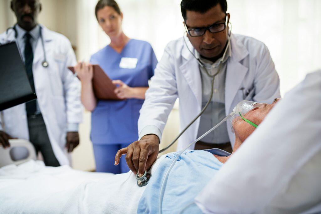 Doctor uses a stethoscope to examine a hospitalized patient receiving oxygen while other medical staff observe in a clinical hospital setting.