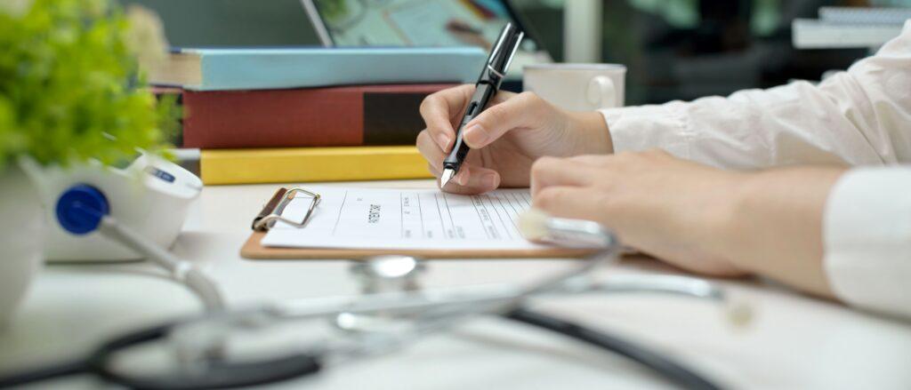 Close-up of a healthcare professional filling out a medical form on a clipboard with a pen, with a stethoscope and medical equipment visible on the desk.
