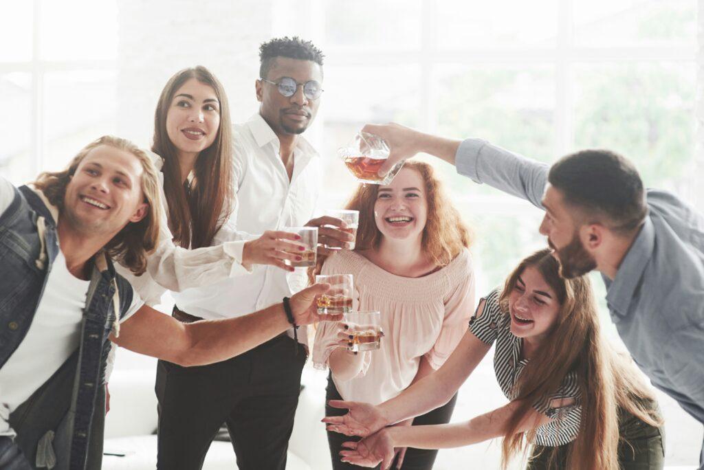 Group of young adults gathered indoors, smiling and laughing as they raise glasses of alcohol in a celebratory toast, with casual clothing, relaxed body language, and bright natural light coming through large windows in the background.
