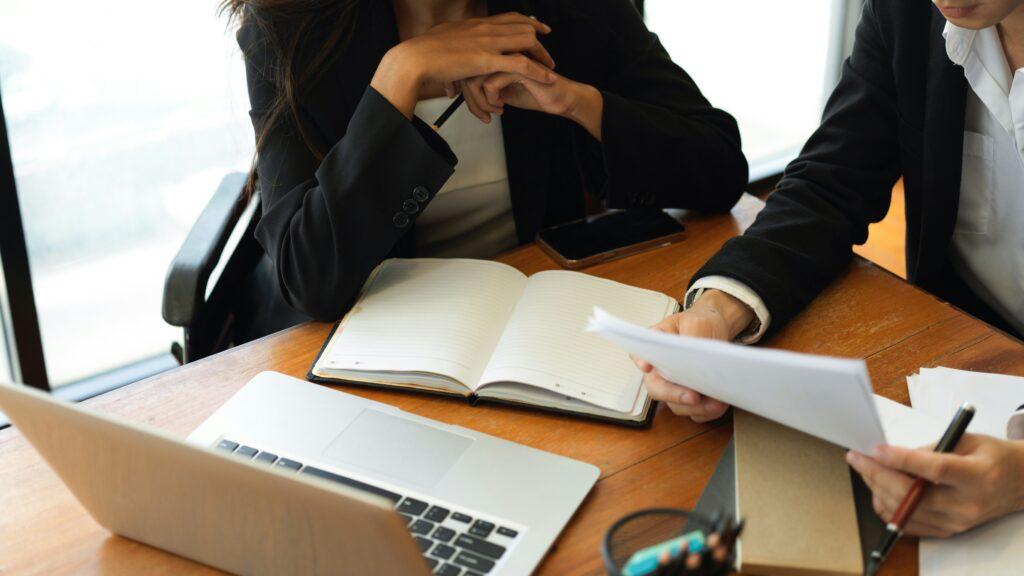 Two professionals in business attire review printed documents at a wooden desk with an open laptop, notebook, and smartphone during a focused office meeting.