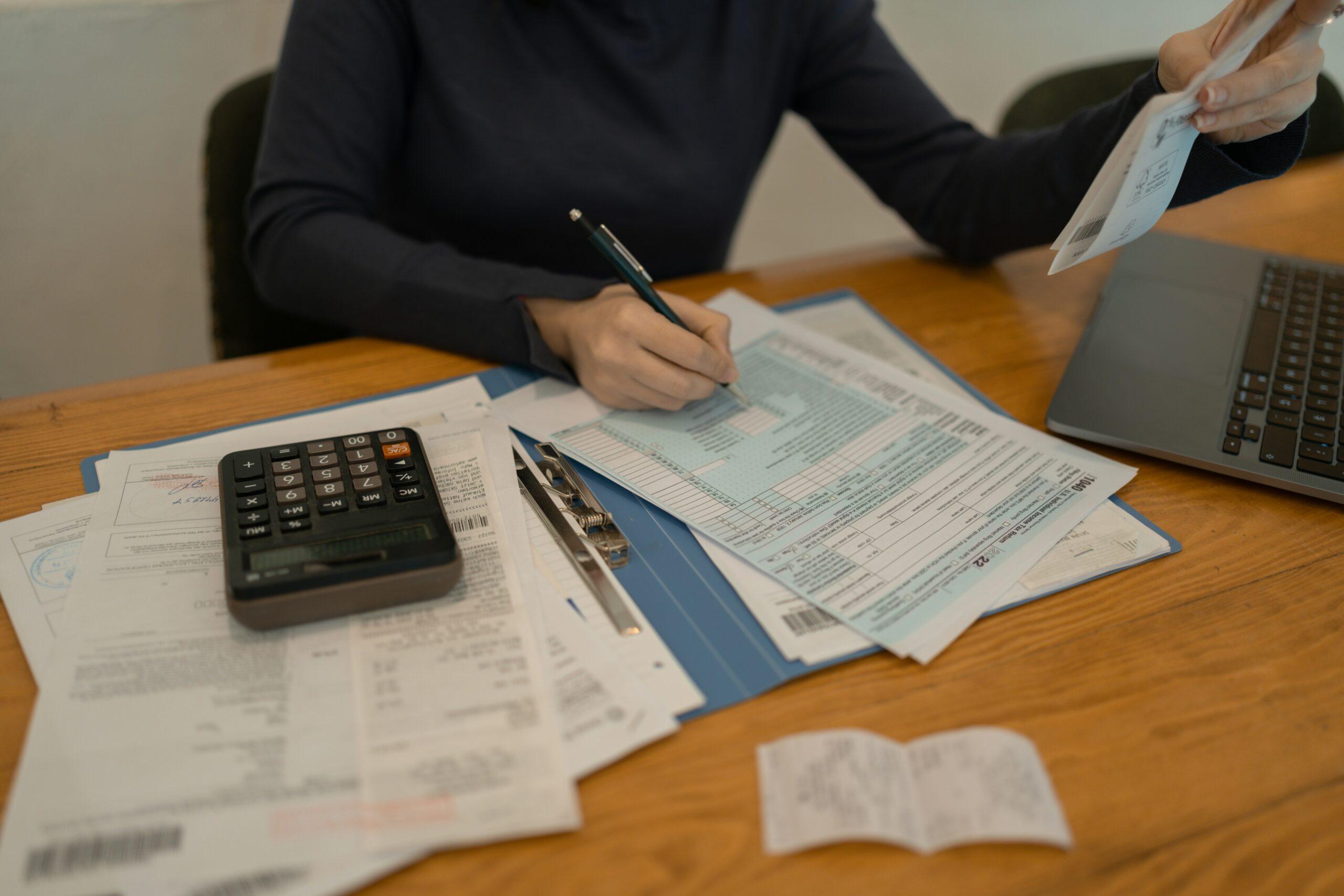 Person seated at a wooden desk filling out official-looking forms with a pen, surrounded by scattered paperwork and receipts, a calculator resting on top of documents, and an open laptop to the side, suggesting careful review and completion of financial, legal, or administrative paperwork.