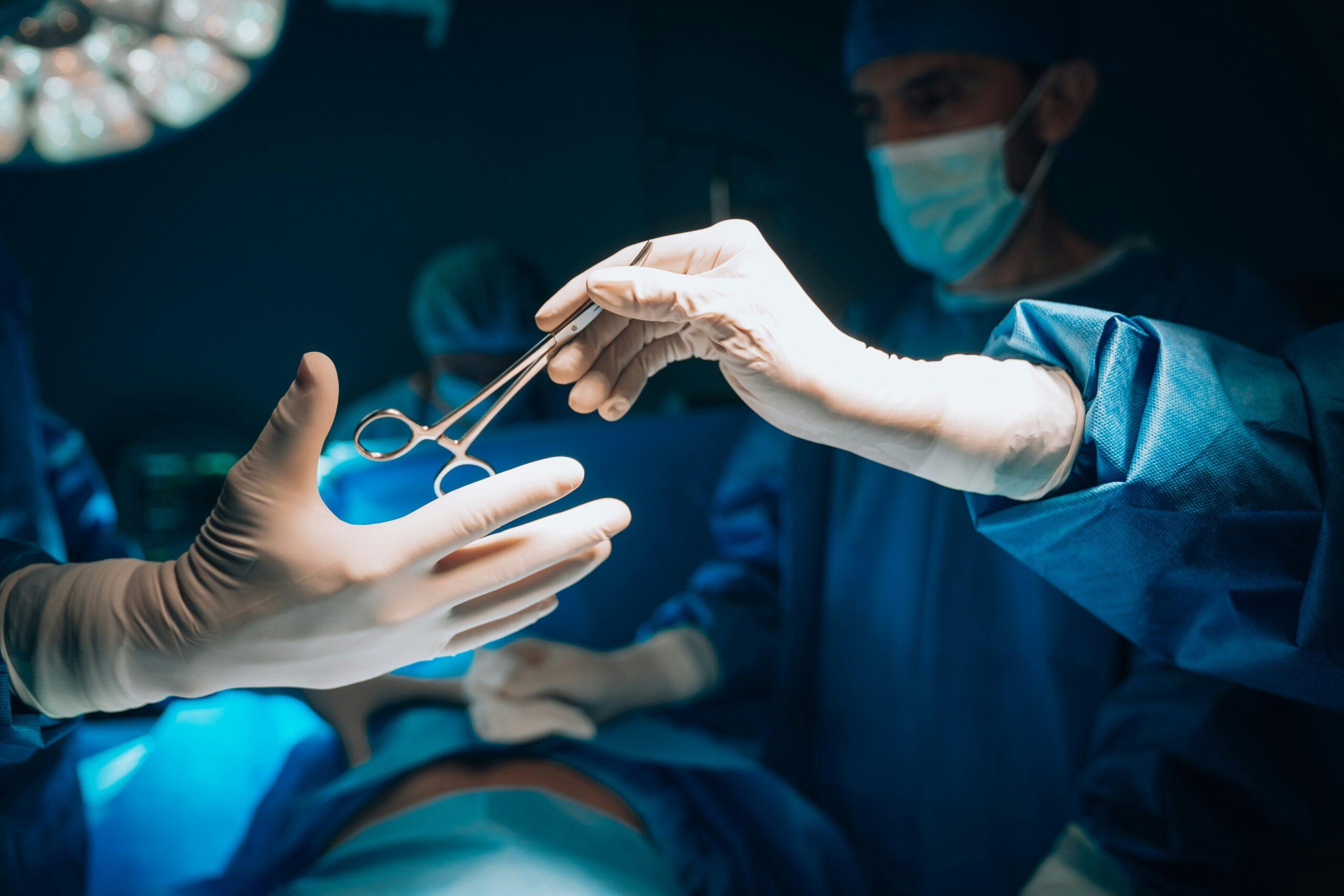 Close-up view inside an operating room showing gloved hands of medical professionals passing a surgical instrument during a procedure, with blue surgical gowns, sterile gloves, and face masks visible under bright operating lights, while a patient lies draped in surgical sheets in the background, creating a focused, sterile, and high-intensity clinical atmosphere.