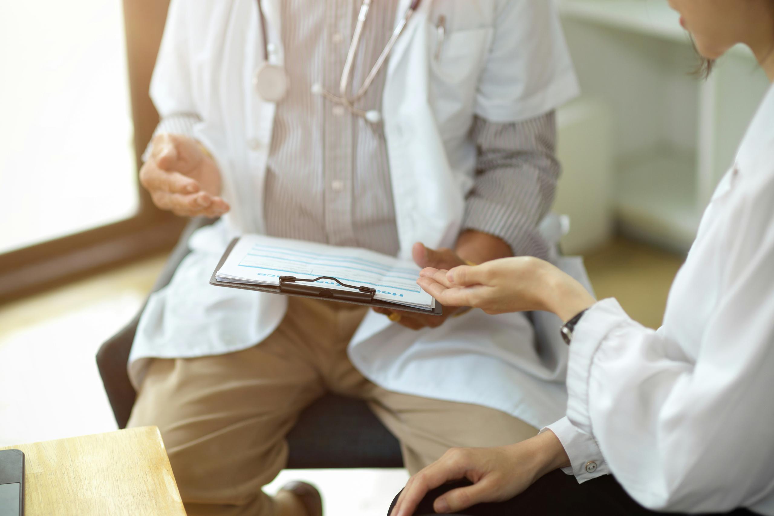 Two healthcare professionals seated indoors reviewing a clipboard with medical notes, both wearing white lab coats, gesturing with their hands as they discuss information in a bright, clinical office setting.