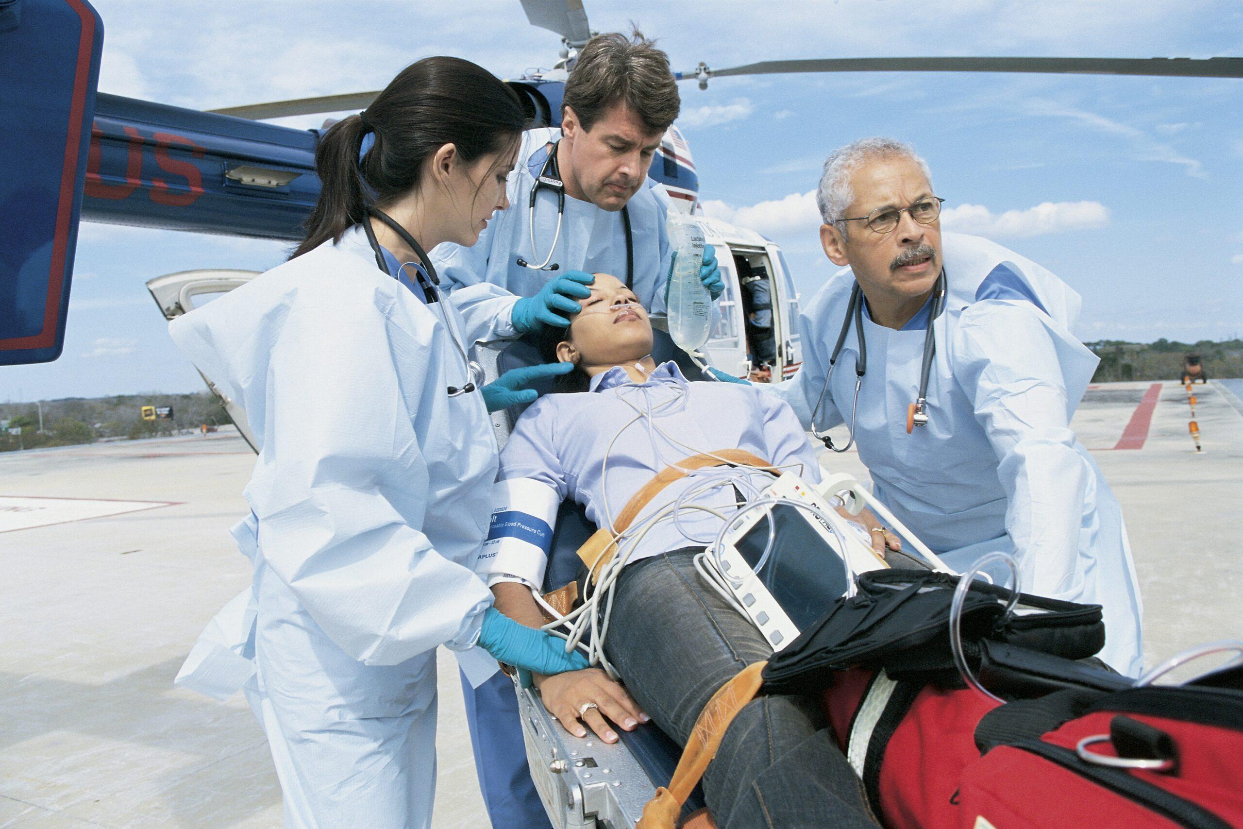 Emergency medical team treating a patient on a stretcher beside a helicopter, with paramedics in protective gowns and gloves administering care and monitoring vital signs on a helipad under a clear sky.