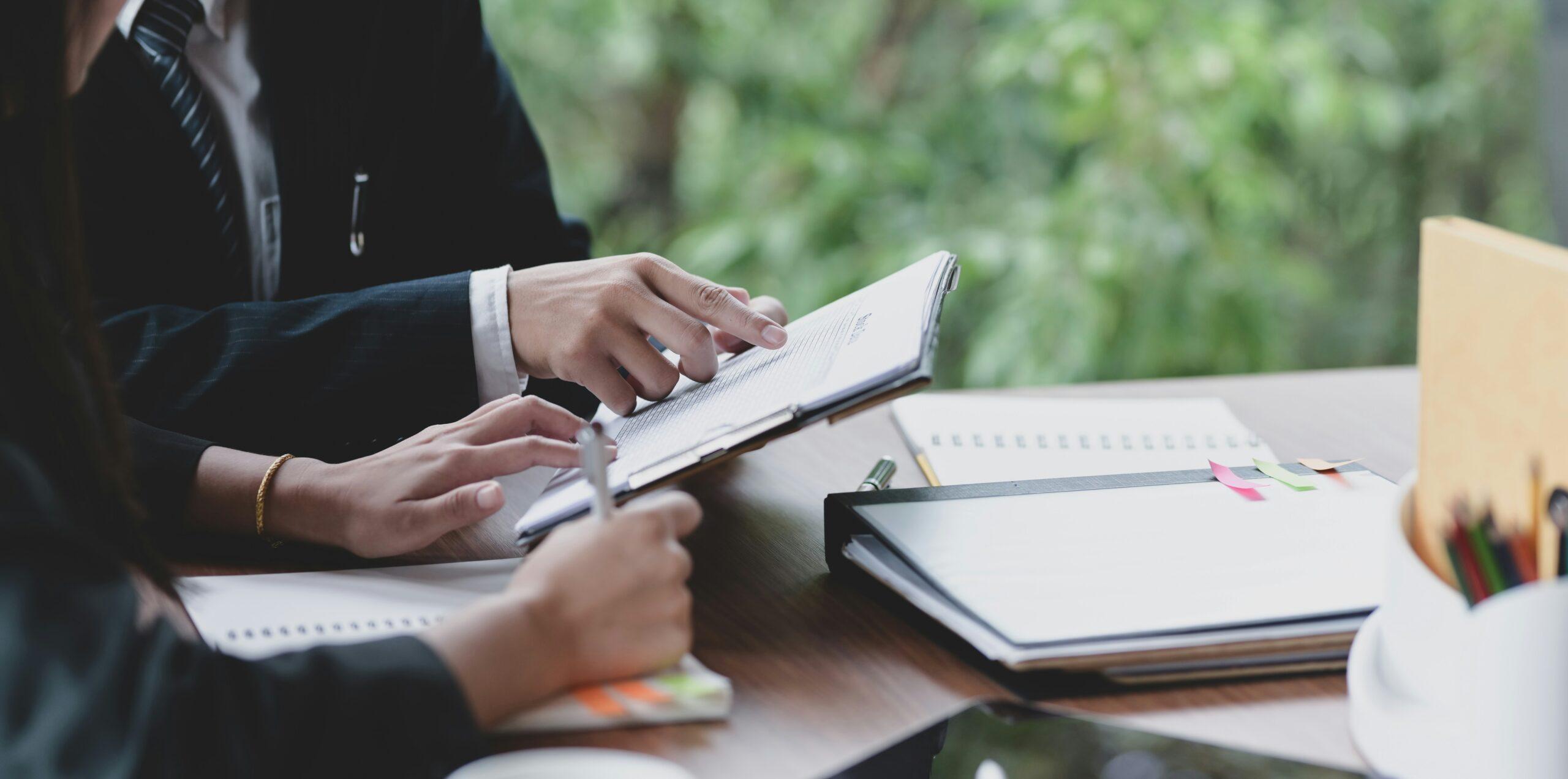 Close-up of two professionals seated at a table reviewing documents, with one person pointing to a specific section on a clipboard while the other takes notes, surrounded by folders, notebooks, and office supplies, with greenery visible through a window in the background.