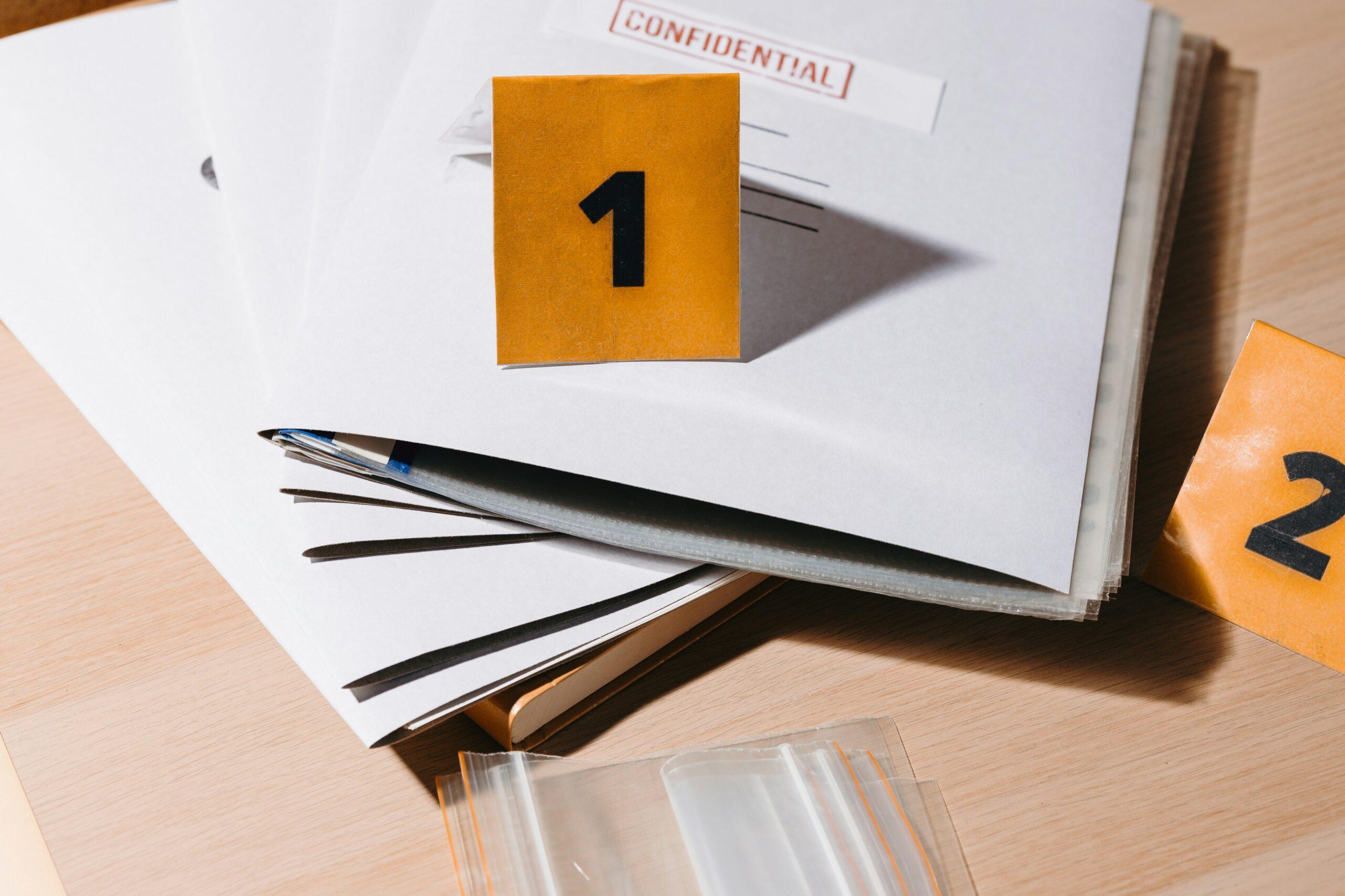 A stack of paper documents spread across a wooden desk, partially labeled “CONFIDENTIAL,” with orange evidence markers showing the numbers one and two placed on top, a pen lying diagonally across the papers, and clear plastic evidence bags nearby, suggesting organized case files or legal evidence under review.