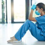 Exhausted healthcare worker in medical scrubs and face mask sitting on the floor with head in hand inside a hospital or clinic hallway