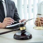 Man holding a pen over papers on a clipboard beside a gavel and scales of justice, with another person's clasped hands nearby