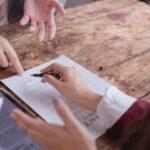 Person pointing to the signature line of a contract as another person signs it with a pen